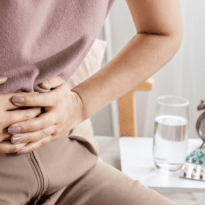 A person holding their abdomen in discomfort sits beside a table with a glass of water, Enim medication, and an alarm clock.