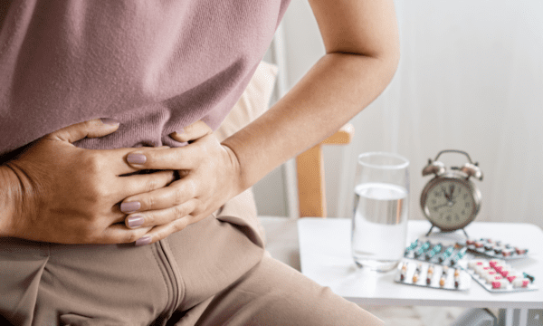 A person holding their abdomen in discomfort sits beside a table with a glass of water, Enim medication, and an alarm clock.