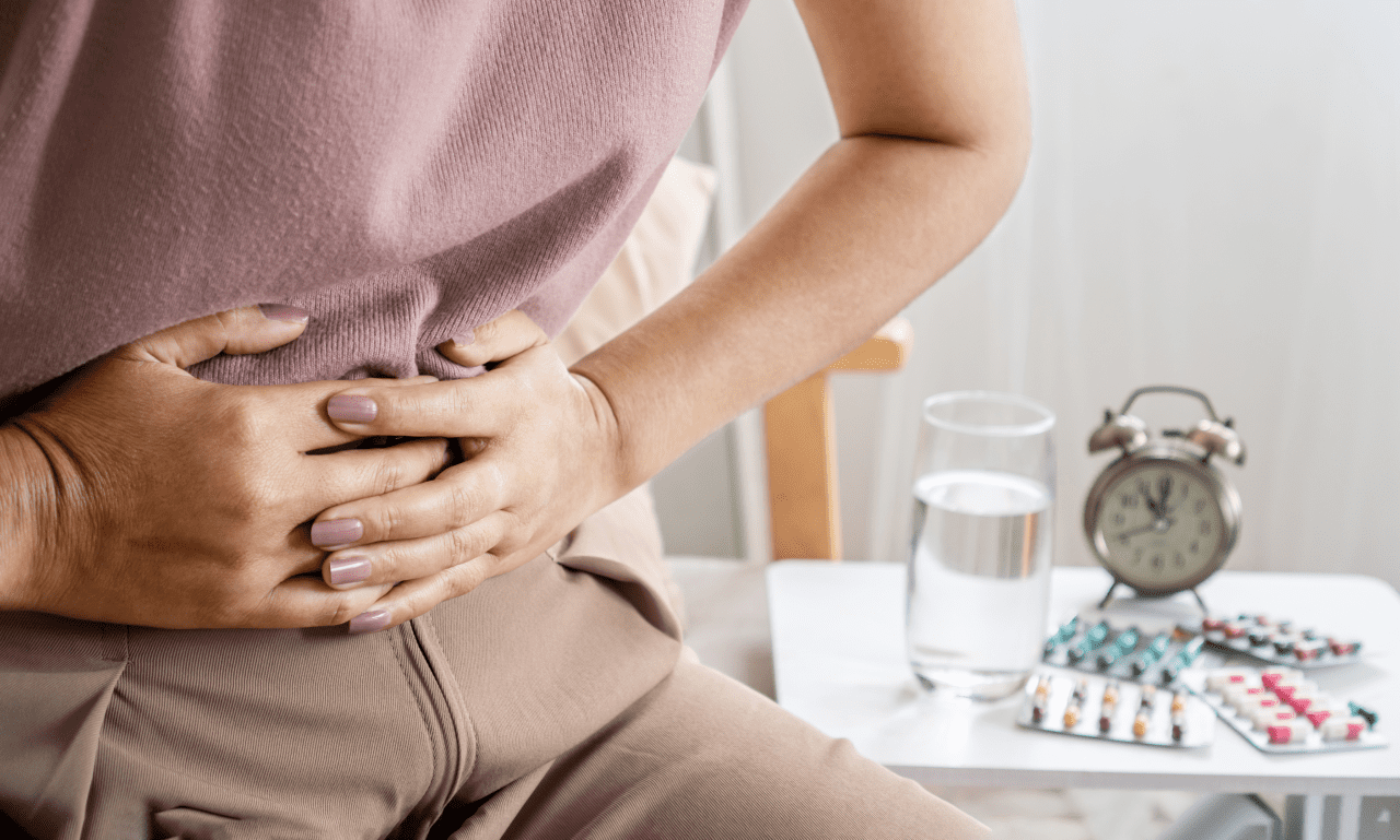 A person holding their abdomen in discomfort sits beside a table with a glass of water, Enim medication, and an alarm clock.